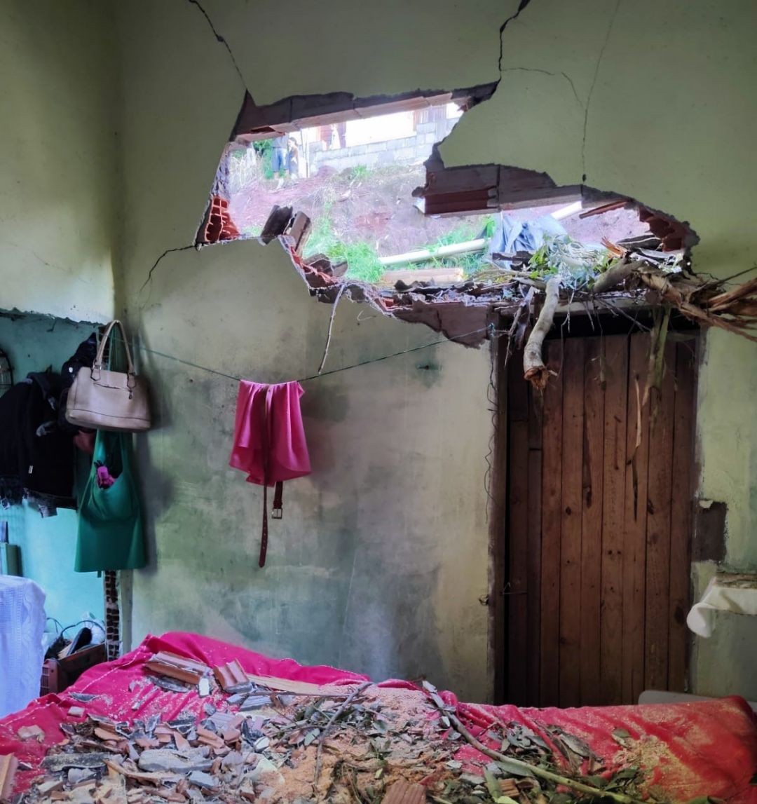 Parede de residência em Ibatiba destruída após queda de eucalipto, com telhado rompido e entulho sobre a cama.