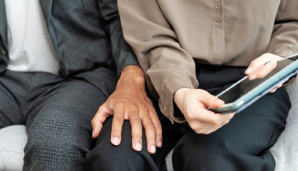 Mão de um homem tocando a perna de uma mulher sentada ao lado dele, enquanto ela segura um celular, representando situação de assédio ou importunação sexual.