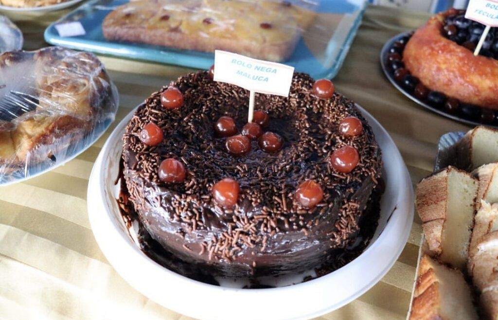 Bolo de chocolate com cobertura de chocolate granulado e cerejas no topo, exposto sobre uma mesa ao lado de outros bolos e pães, indicando produção artesanal de confeitaria.