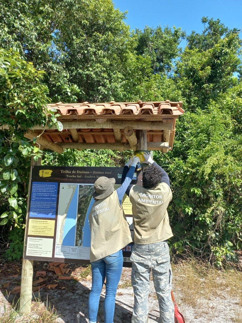 Monitores ambientais instalam placa informativa em trilha do Parque Estadual Cachoeira da Fumaça, cercada por vegetação nativa.