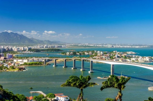 Vista panorâmica de Vitória, com a Terceira Ponte ligando a capital a Vila Velha sobre o mar, prédios ao fundo, áreas verdes e céu azul em dia ensolarado.