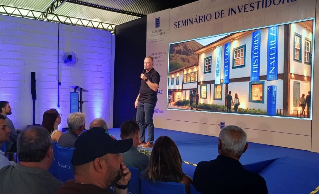 O governador Renato Casagrande fala aos presentes ao Seminário de Investidores do Complexo Turístico Cultural e Ambiental Fazenda Santa Maria, em Muniz Freire, tendo a imagem do complexo em painel atrás dele.