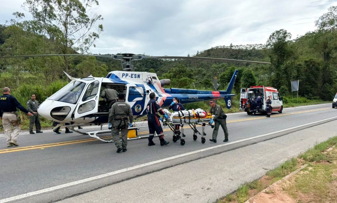 Helicóptero do Notaer pousado na BR-262 enquanto equipes do Corpo de Bombeiros e do SAMU realizam o resgate de vítima em maca após acidente entre carro e caminhão.