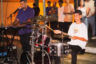 Jovem toca bateria durante apresentação musical no evento Despertai Ibitirama, com público acompanhando ao fundo.