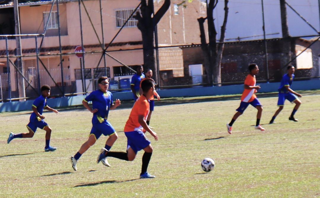 Jovens atletas em campo durante uma partida de futebol de base. Ao centro, um jogador com uniforme laranja e azul conduz a bola, sendo acompanhado por outros atletas em um campo gramado sob a luz do dia.