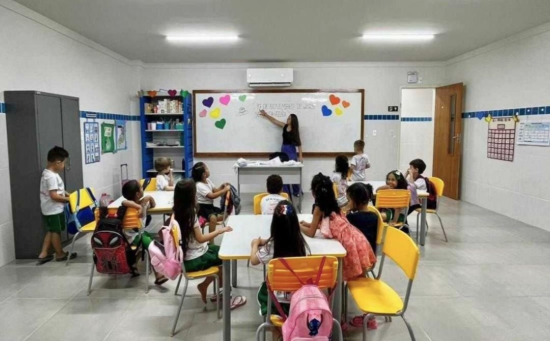 Sala de aula da rede municipal de Muniz Freire com professora em atividade no quadro e alunos sentados em mesas, representando o ensino público infantil e a valorização da educação.
