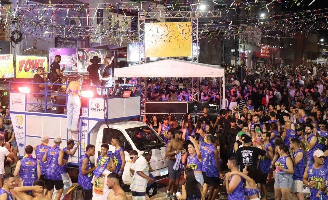 Multidão acompanha trio elétrico durante carnaval noturno em Dores do Rio Preto, com foliões usando abadás e blocos nas ruas iluminadas.