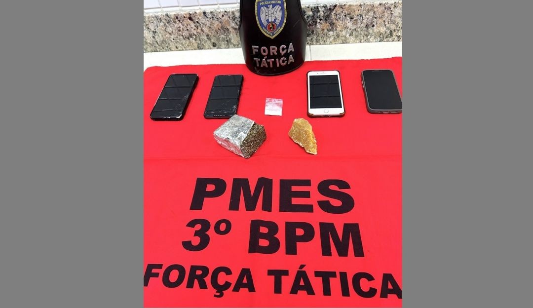 Imagem mostra mesa coberta com pano vermelho identificado como “PMES 3º BPM Força Tática”, onde estão expostos um tablete de maconha, uma pedra grande de crack, um papelote de cocaína, uma porção de maconha e quatro aparelhos celulares apreendidos durante ação policial em Alegre.