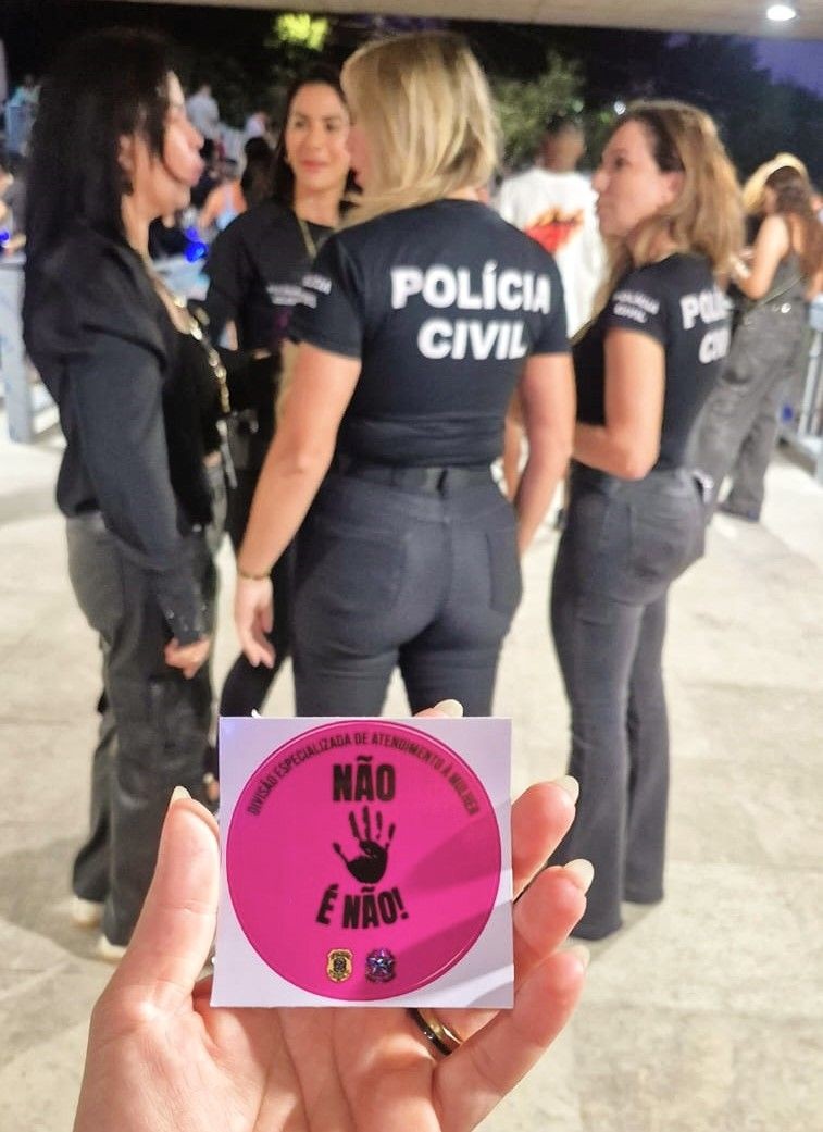 Agentes da Polícia Civil usando camisetas com a inscrição “Polícia Civil” durante operação no Carnaval; em primeiro plano, adesivo rosa com a frase “Não é Não”, símbolo da campanha de combate à importunação sexual.