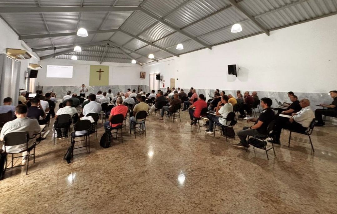 Amplo salão de reuniões com teto metálico e luminárias pendentes. Vários homens (padres e religiosos) estão sentados em cadeiras escolares, de costas para a câmera, assistindo a uma palestra ao fundo do salão, onde há um crucifixo na parede e uma tela de projeção.
