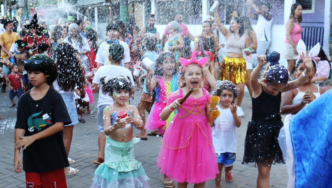 Crianças do Bloco Curumim desfilando pelas ruas de Patrimônio da Penha, no Carnaval 2026 de Divino de São Lourenço, fantasiadas, sob uma chuva de confete e espuma.