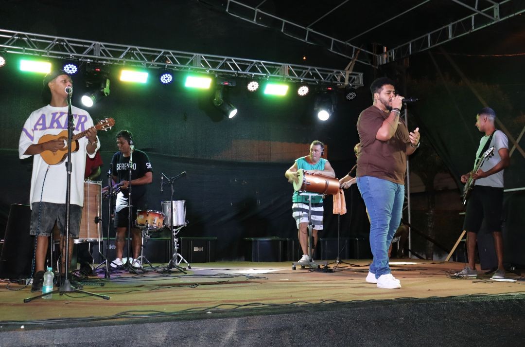 Banda se apresenta no palco montado na Praça do CRAS durante o Carnaval 2026 em Divino de São Lourenço, com cantor ao microfone, músicos tocando instrumentos de percussão e cordas e iluminação colorida ao fundo.