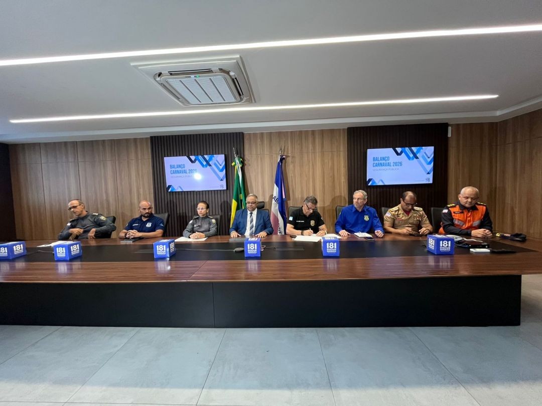 Autoridades da Segurança Pública do Espírito Santo participam de coletiva de imprensa sobre o Balanço do Carnaval 2026. Sete representantes das forças de segurança estão sentados atrás de uma mesa de reuniões, com bandeiras ao fundo e dois telões exibindo a identidade visual do evento.