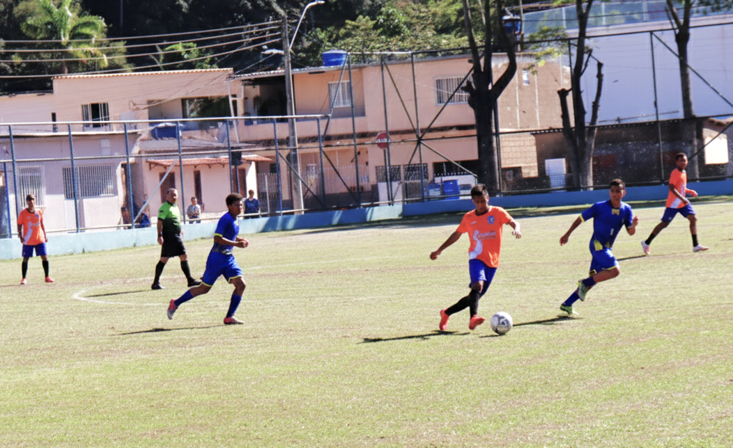 Jogadores disputam partida de futebol no campo de Dores do Rio Preto-ES, tendo três com camisa laranja e outros dois com uniforme azul, além do arbitro de verde e preto que acompanha a jogada.