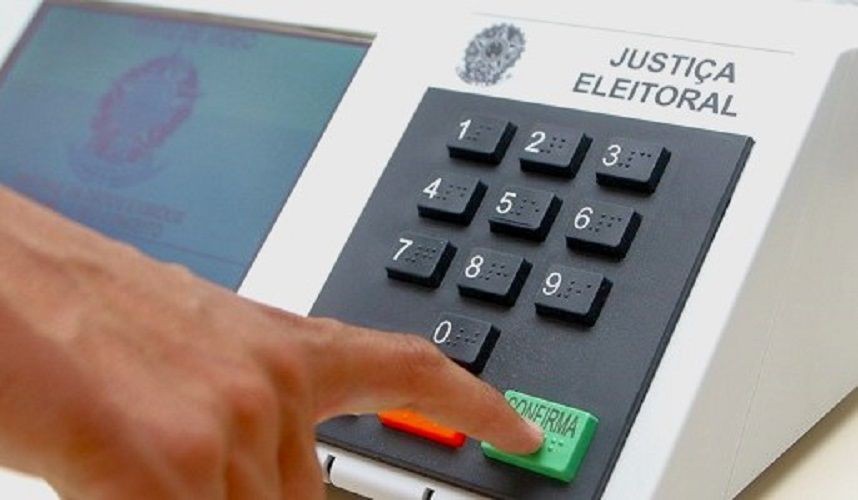Imagem mostra uma urna eletrônica da Justiça Eleitoral brasileira, com destaque para o teclado numérico e o botão verde “Confirma”, sendo pressionado por um eleitor.