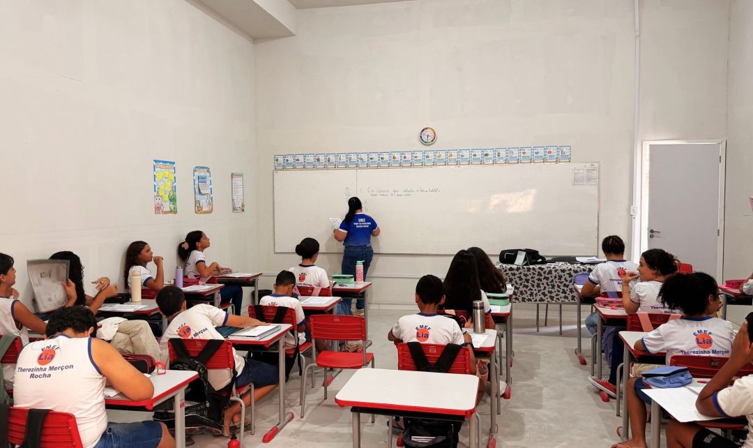 Professora ministra aula em sala da rede municipal de ensino de Muniz Freire, com alunos sentados em carteiras vermelhas enquanto acompanham atividade no quadro branco.