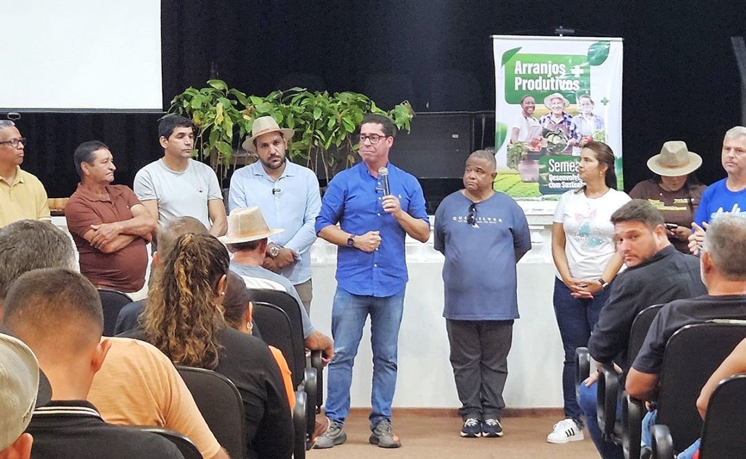 Produtores rurais e autoridades participam de cerimônia de entrega de mudas do Projeto Arranjos Produtivos, em auditório, com banner do programa ao fundo