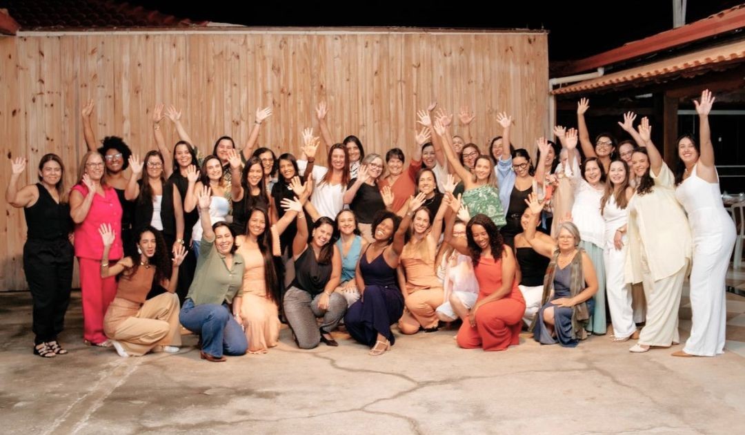 Grupo de 40 mulheres sorridentes e acenando, com braços levantados, durante foto oficial do evento Café com Ideias em um ambiente amplo com parede de madeira ao fundo.