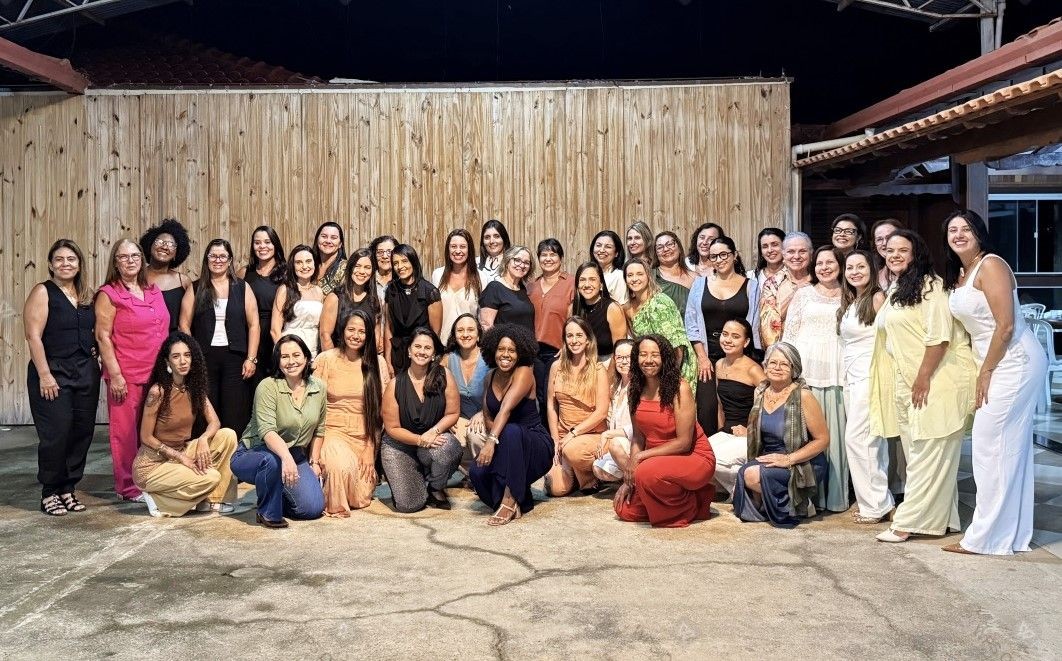 Grupo de cerca de 40 mulheres sorridentes posando para foto oficial do evento Café com Ideias em um ambiente amplo com parede de madeira ao fundo.
