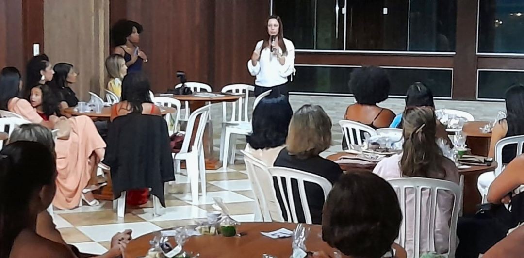 Cyntia Grillo, de blusa branca, falando ao microfone para uma plateia de mulheres sentadas em mesas redondas durante o evento em Iúna.