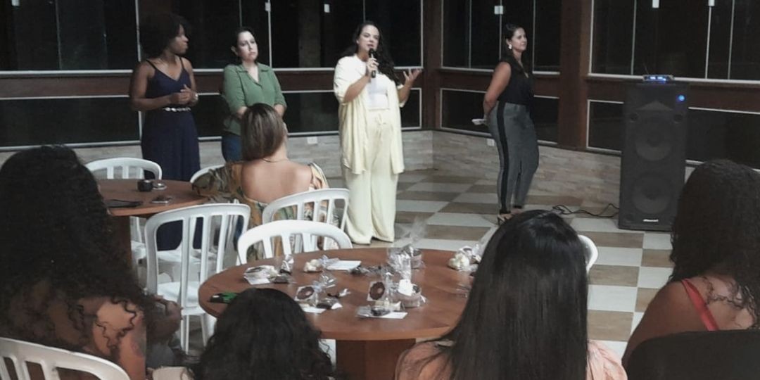 Quatro mulheres, da diretoria do movimento, a frente de outras mulheres sentadas em mesas redondas que estão ouvindo a fala de uma delas, durante o Café com Ideias.