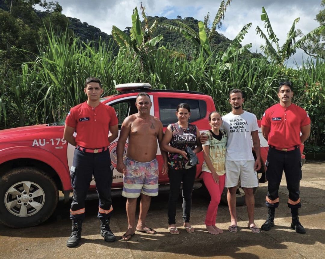 Foto dos dois irmãos que estavam perdidos no Parque Nacional do Caparaó, com dois bombeiros da equipe de resgate e um homem sem camisa e uma mulher moradores da residência em Santa Marta, Ibitirama, na frente de uma viatura do Corpo de Bombeiros e ao fundo vegetação típica da zona rural e da Serra do Caparaó.