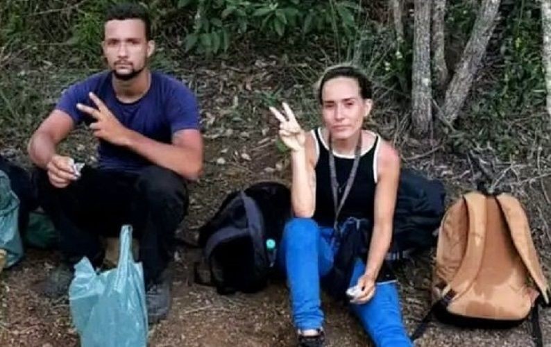 Casal de irmãos sentado em área de mata, com mochilas ao lado, fazendo sinal de positivo após serem encontrados na região do Caparaó.