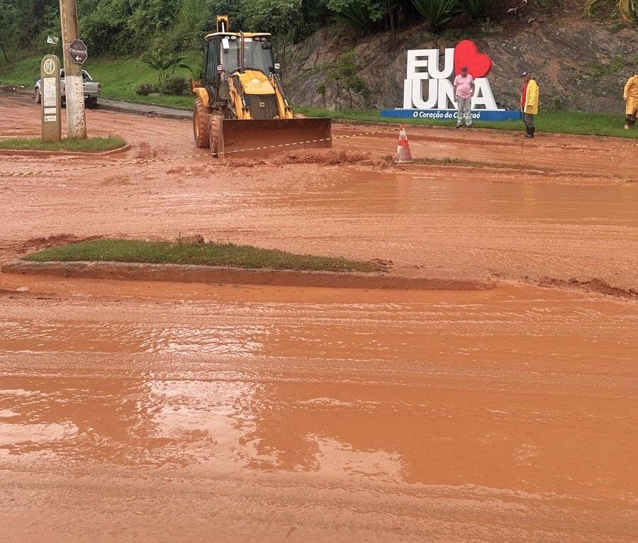 Máquina retroescavadeira realiza a retirada de lama em via urbana de Iúna após forte chuva; ao fundo, o letreiro “Eu ❤️ Iúna – O Coração do Caparaó” aparece em meio ao cenário de ruas cobertas por barro.