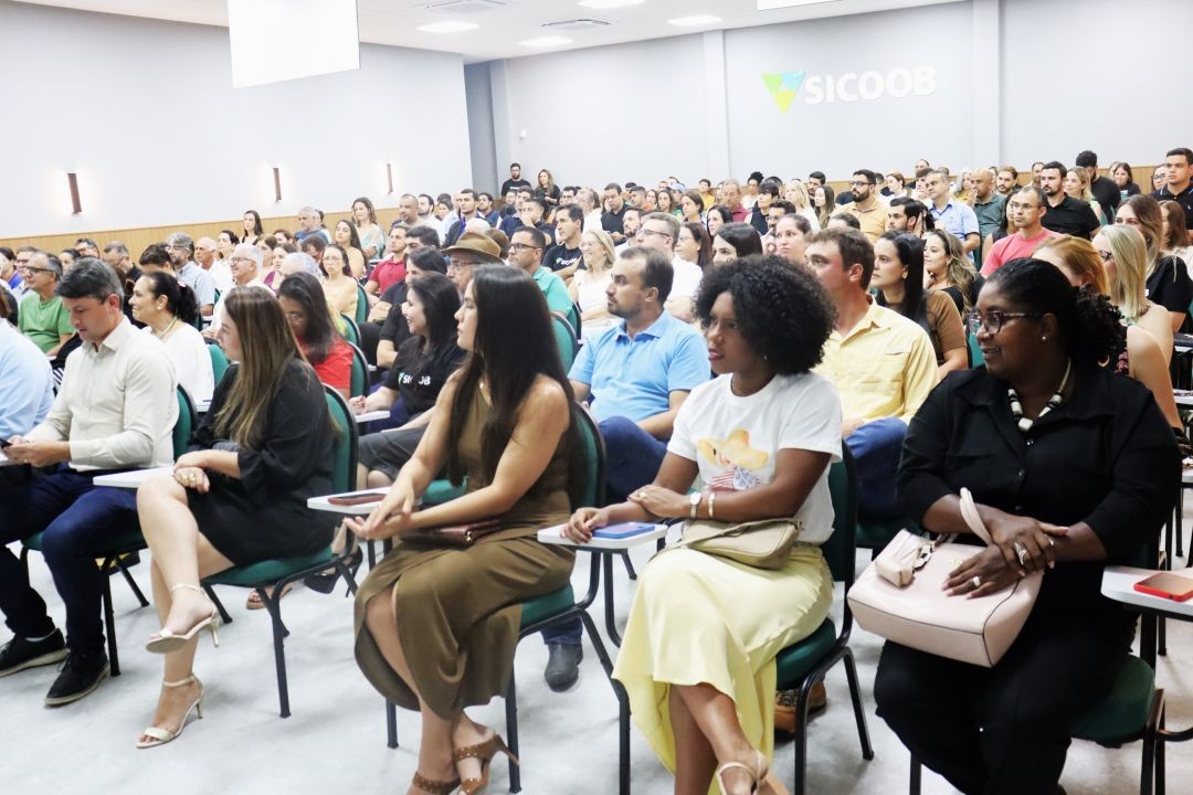 Vista lateral de um auditório cheio na agência do Sicoob Iúna durante a palestra sobre Reforma Tributária. Homens e mulheres de diversas idades estão sentados em cadeiras verdes com pranchetas acopladas, ouvindo uma palestra. Ao fundo, na parede cinza, está o logotipo iluminado do Sicoob.