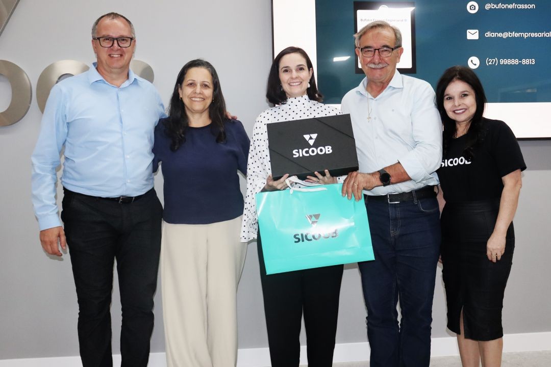 Cinco pessoas posam para uma foto oficial em frente ao logotipo do Sicoob. Ao centro, a palestrante Dra. Diane Espíndola segura uma caixa de presente e uma sacola personalizada da cooperativa. Ela veste uma blusa branca com estampa de poá preto. Ao seu lado, quatro representantes do Sicoob (três homens e uma mulher) sorriem. No canto superior direito, há uma tela com informações de contato.