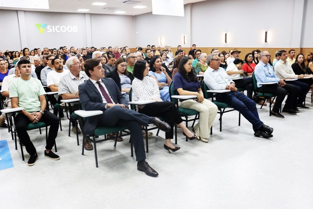 Vista lateral de um auditório lotado na agência do Sicoob Iúna durante a palestra sobre Reforma Tributária. O público, composto por homens e mulheres de diversas idades, está sentado em cadeiras verdes com pranchetas acopladas, ouvindo atentamente. Na primeira fileira, destacam-se a Dra. Diane Espíndola e outros convidados. Ao fundo, na parede cinza, vê-se o logotipo iluminado do Sicoob.
