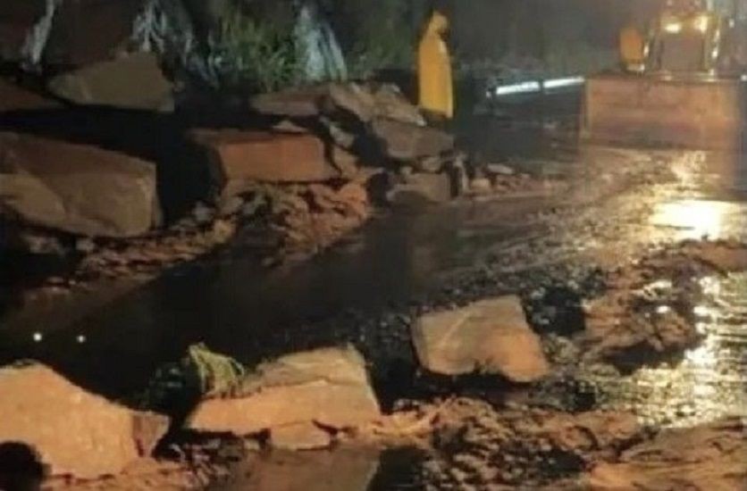 Imagem noturna mostra grandes rochas obstruindo a pista da rodovia ES-181 em Alegre, com uma máquina retroescavadeira e um técnico da Defesa Civil de capa amarela ao fundo iniciando os trabalhos de sinalização.