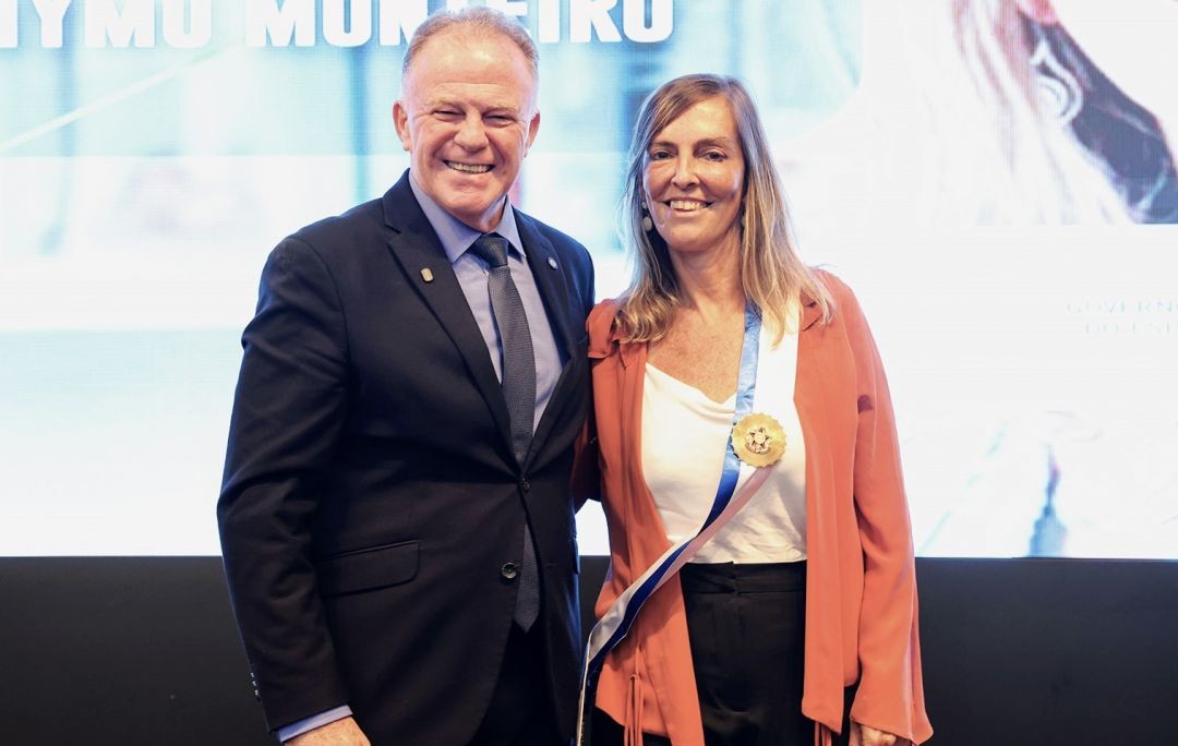 O governador Renato Casagrande, homem de pele clara e terno azul marinho, sorri ao lado da Dra. Tatiana Sampaio. Ela veste uma blusa branca, blazer terracota e ostenta a faixa azul e branca com a medalha da Comenda Jerônymo Monteiro no peito. Ao fundo, um telão exibe o nome do evento.