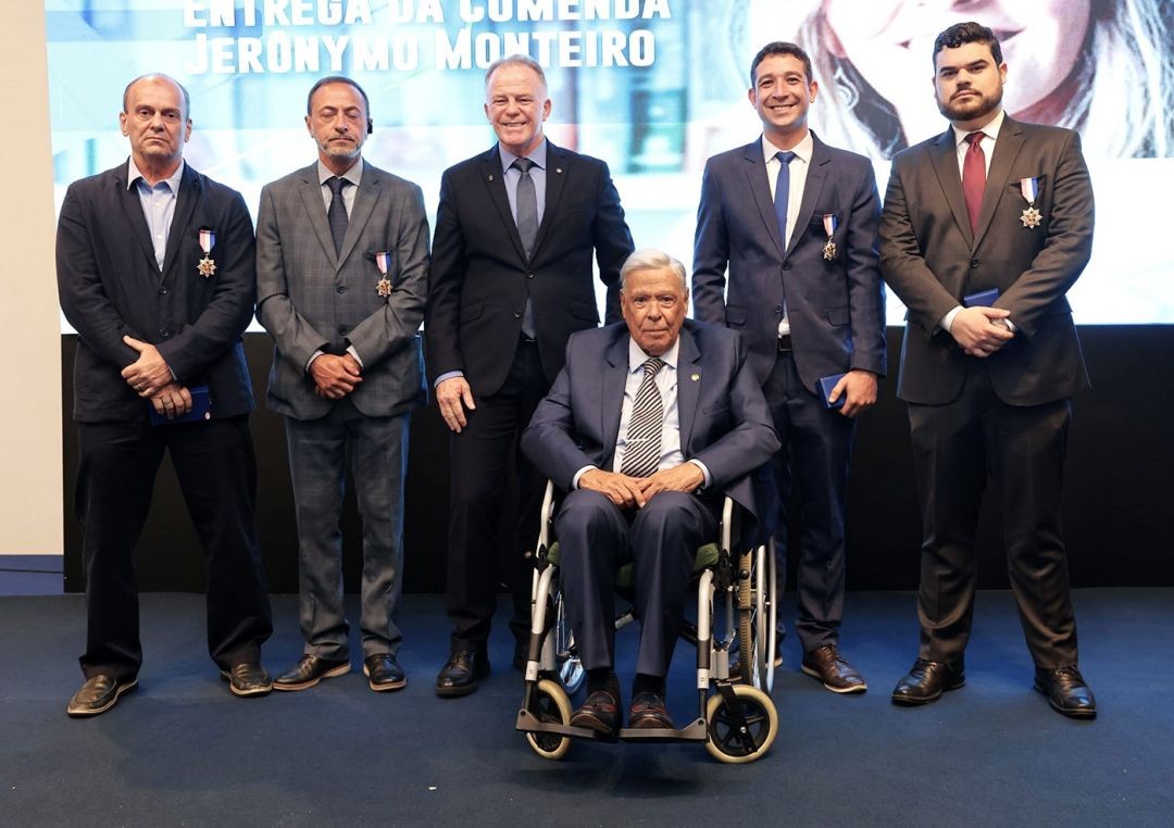Foto oficial do grupo de homenageados no Palácio Anchieta. Ao centro, um senhor em cadeira de rodas usa terno e gravata listrada. Atrás dele, em pé, estão o governador Renato Casagrande e outros quatro homenageados, incluindo Mitter Mayer. Todos vestem trajes formais e exibem as medalhas da Ordem Cavaleiro fixadas nos paletós.