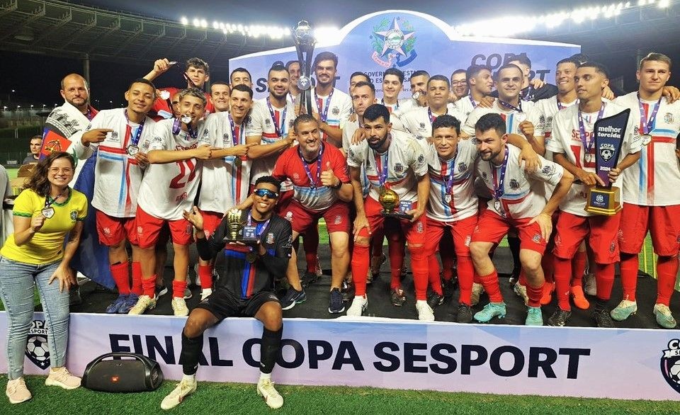 Jogadores do time campeão de Iúna posam para foto segurando troféus e medalhas após a final da Copa Sesport, em campo iluminado à noite, com faixa “Final Copa Sesport” ao fundo.