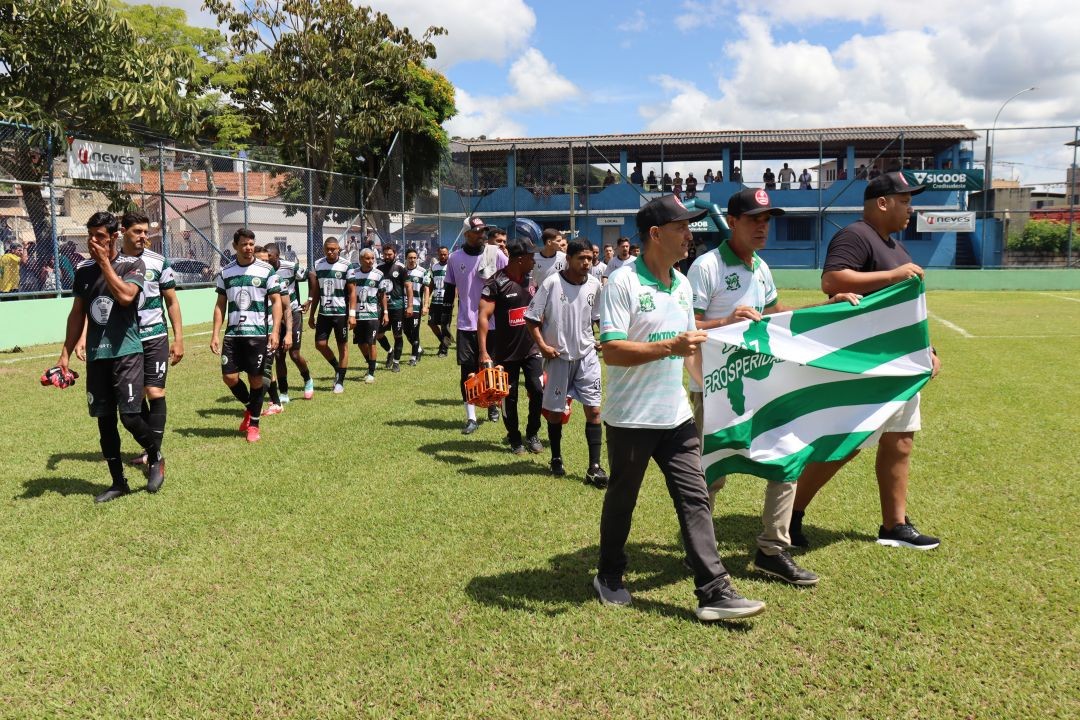 Equipes entram em campo no estádio municipal de Dores do Rio Preto durante a abertura do 1º Campeonato Sicoob Credisudeste, com jogadores à frente segurando bandeira verde e branca.