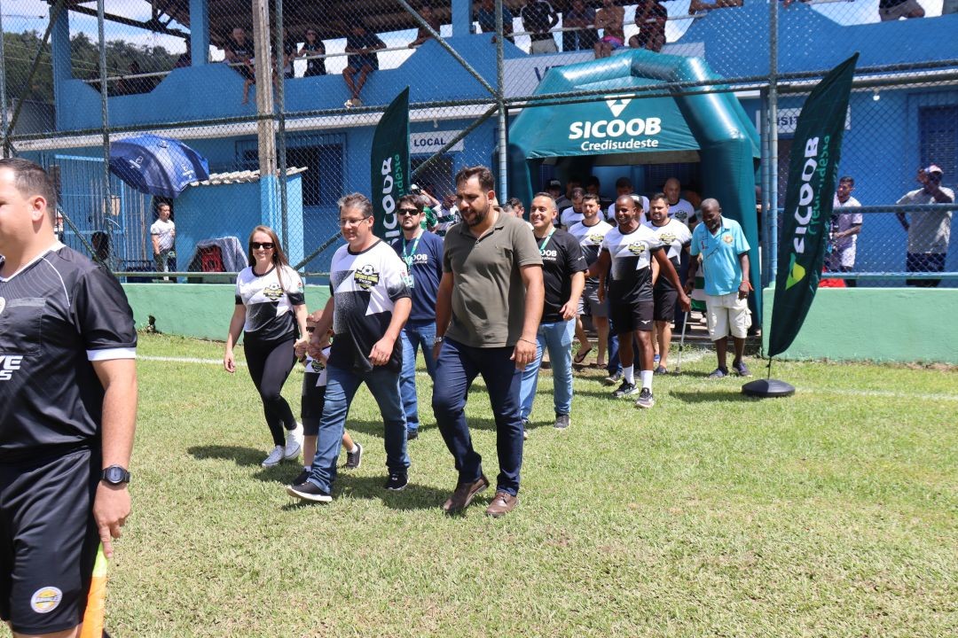 Prefeito de Dores do Rio Preto, primeira-dama e demais autoridades entram em campo pelo túnel inflável do Sicoob Credisudeste durante a abertura do campeonato, acompanhados por organizadores e participantes, com torcedores nas arquibancadas ao fundo.