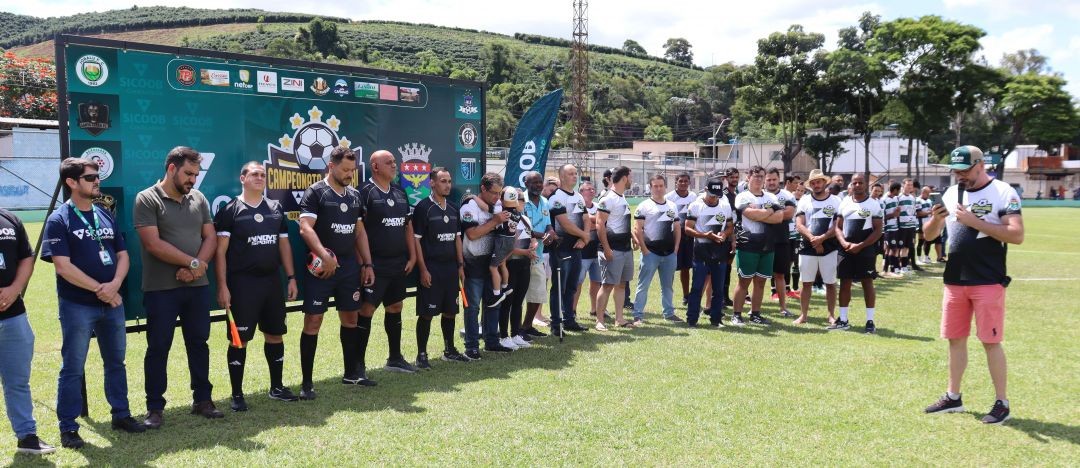 Autoridades municipais, organizadores, representantes do Sicoob Credisudeste e equipes de arbitragem perfilados no gramado durante a cerimônia oficial de abertura do 1º Campeonato Sicoob Credisudeste, em Dores do Rio Preto, com painel oficial da competição ao fundo e público nas arquibancadas.