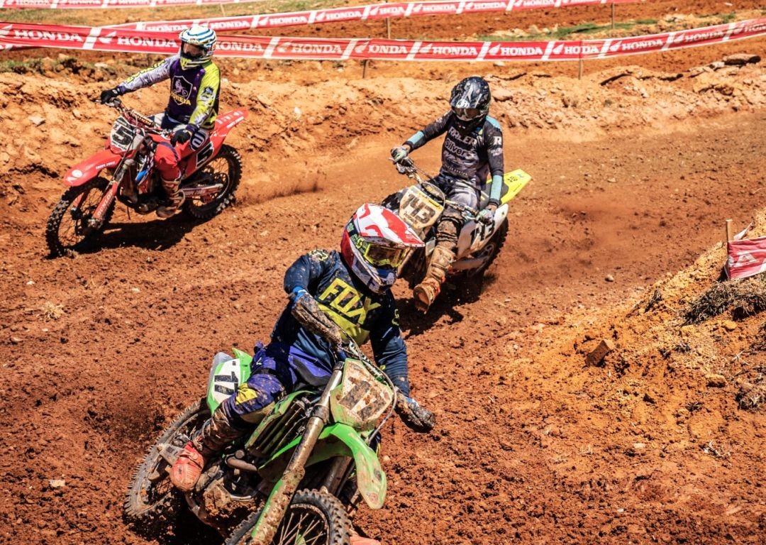 Três pilotos de motocross disputam posição em um trecho de terra batida. Em destaque, na frente, um piloto com uniforme da Fox (azul e amarelo) e capacete branco e vermelho pilota uma moto Kawasaki verde. Logo atrás, outros dois competidores em motos vermelha e branca mantêm a perseguição em alta velocidade.