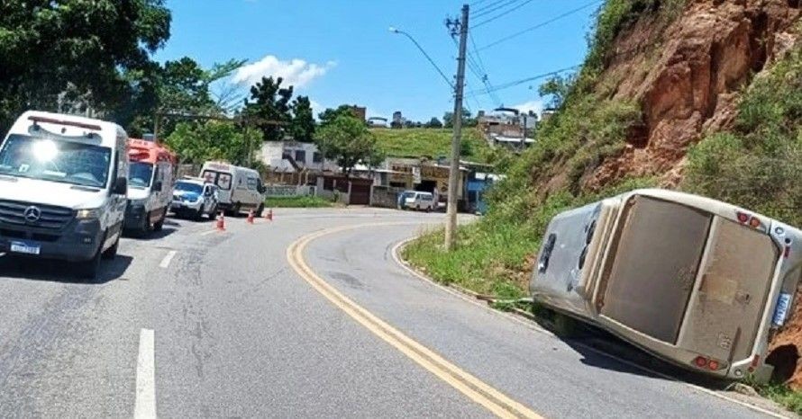 Imagem do micro-ônibus da Secretaria de Saúde de Iúna no dia do acidente, tombado à margem da rodovia BR-482, na descida da Serra de Alegre, com ambulâncias e viatura da Polícia Militar parados do outro lado da rodovia, além de casas e outros veículos ao fundo, em curva.