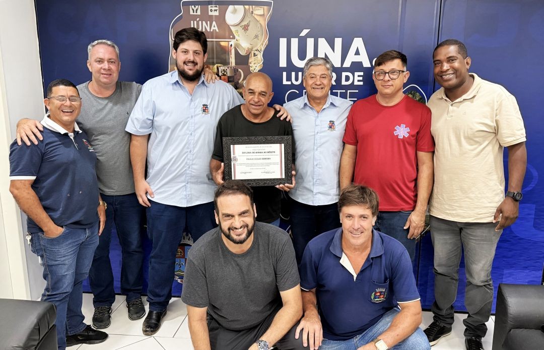 Autoridades municipais e colegas de trabalho posam ao lado do motorista homenageado em frente a painel institucional da Prefeitura de Iúna; ele segura o diploma de Honra ao Mérito.