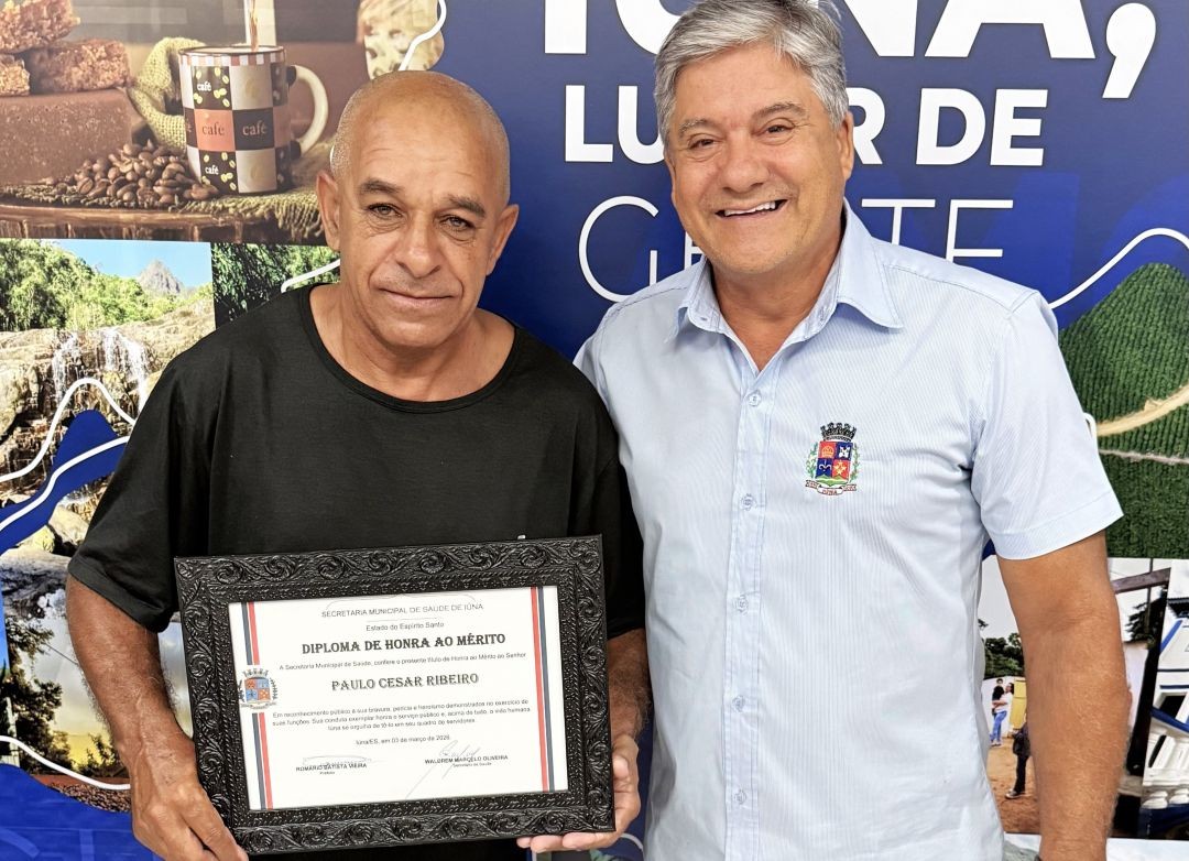 O motorista Paulo Cesar Ribeiro, um homem de pele morena e camiseta preta, segura o certificado emoldurado. Ao seu lado, o Prefeito de Iúna, Romário Batista Vieira, veste uma camisa social azul clara com o brasão do município e sorri para a foto. Eles estão à frente de um painel azul com fotos da cidade e a frase parcial 