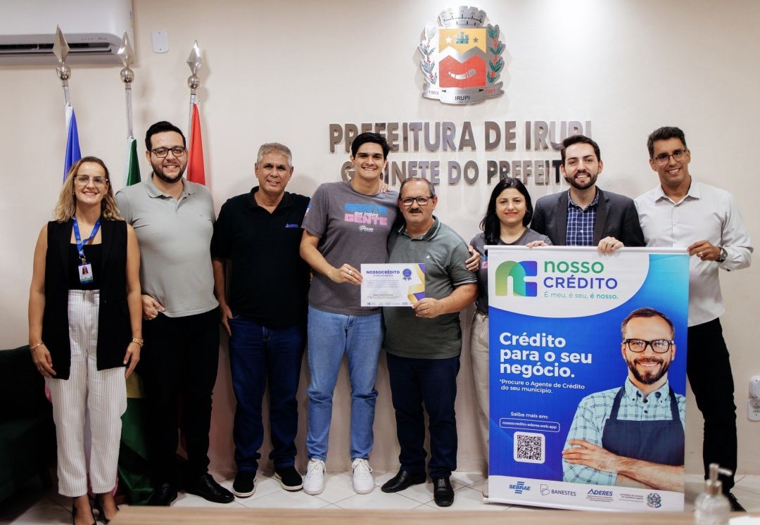 Fotografia em ambiente interno (Gabinete do Prefeito) com o brasão de Irupi ao fundo. Um grupo de sete pessoas (quatro homens e três mulheres) posa sorridente. Ao centro, dois homens seguram um certificado de reconhecimento. À direita, dois homens seguram um banner vertical azul do programa 