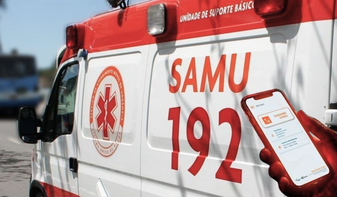 Fotografia lateral de uma ambulância branca e vermelha do SAMU 192. Em destaque, à direita, uma mão segura um smartphone exibindo a interface do aplicativo 