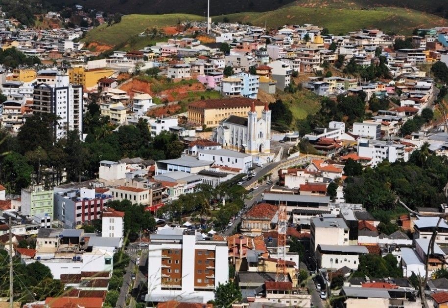 Vista aérea da cidade de Alegre, no sul do Espírito Santo, mostrando a área central com a igreja matriz ao centro, prédios comerciais e residenciais ao redor e morros com casas ao fundo.