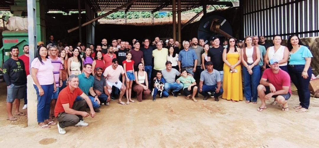 Um grande grupo de pessoas, composto por moradores das comunidades anexadas e autoridades políticas, posa para uma foto oficial dentro de um galpão industrial de beneficiamento de café em Divino de São Lourenço. Ao centro, em pé, destacam-se o prefeito Dudu Queiroz (camisa amarela) e os deputados Marcelo Santos e Fabrício Gandini.