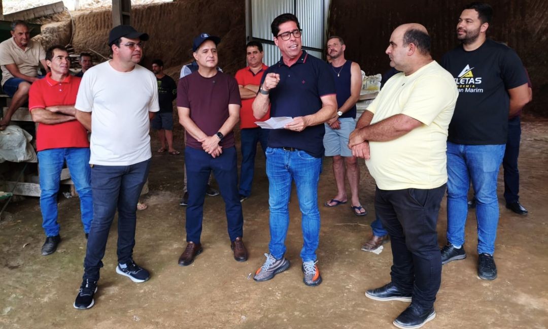 O deputado Marcelo Santos, em pé ao centro, veste camisa polo preta e calça jeans, gesticulando enquanto fala para um grupo de homens em um galpão de processamento de café. Ao seu lado, o prefeito Dudu Queiroz (camisa amarela) e outros representantes acompanham o discurso.