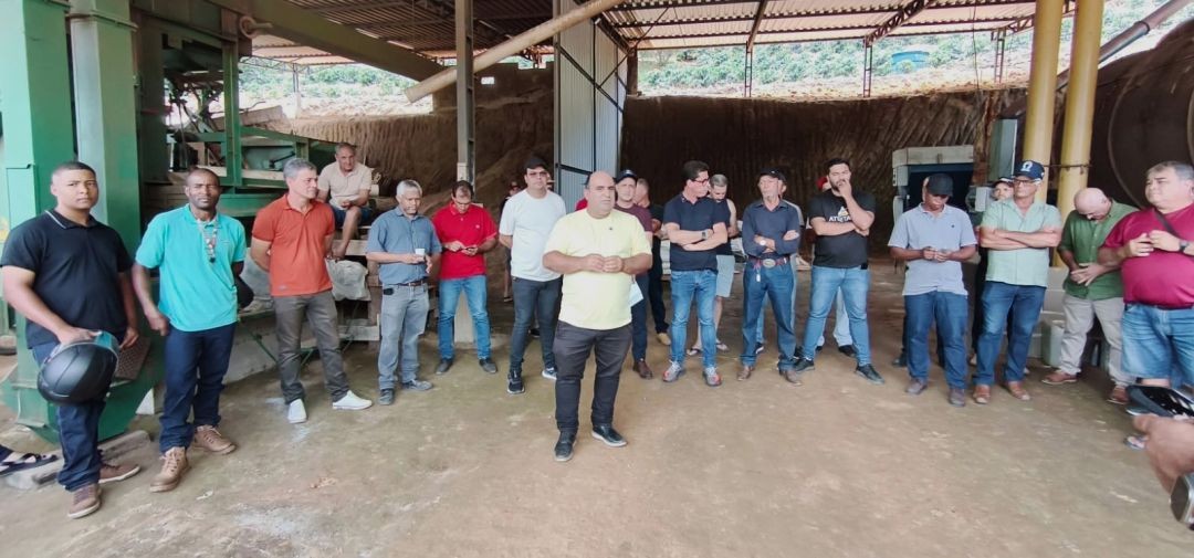 Vista ampla do interior de uma unidade de beneficiamento de café em Divino de São Lourenço. O prefeito Dudu Queiroz, de camisa amarela, fala ao centro para uma comitiva de cerca de 15 pessoas, incluindo autoridades e produtores locais. Ao fundo, veem-se máquinas agrícolas e o terreno escavado.