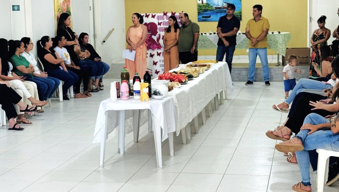 Servidoras de Divino de São Lourenço sentadas em cadeiras em volta de uma mesa com o café da manhã, enquanto ouvem a mensagem de autoridades presentes.