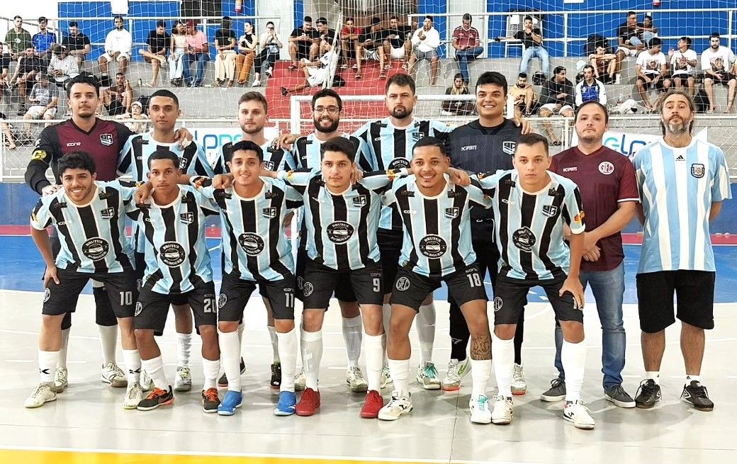 Foto oficial do time do Grêmio. Jogadores posam para a foto em um ginásio poliesportivo. Eles vestem uniforme listrado em azul claro, branco e preto, com o patrocínio 